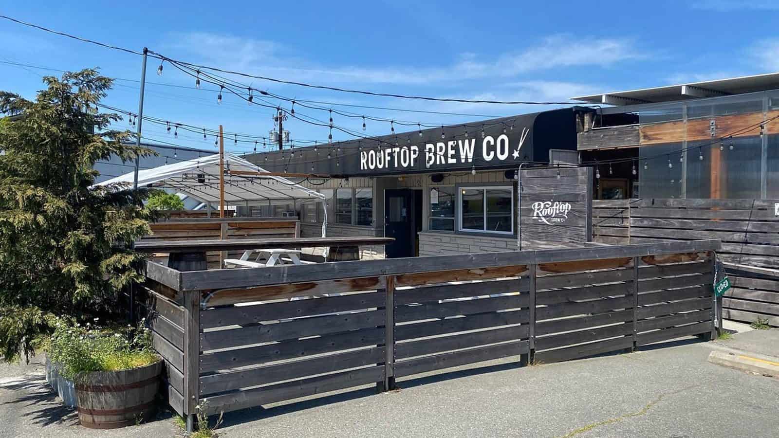 Rooftop_Patio_1600x900 Outdoor view of a wooden rooftop brewery with string lights, a fenced patio area, and a sign reading "Rooftop Brew Co." on a sunny day. After recovering from a devastating fire, Rooftop Brew now stands as a testament to resilience and community spirit.