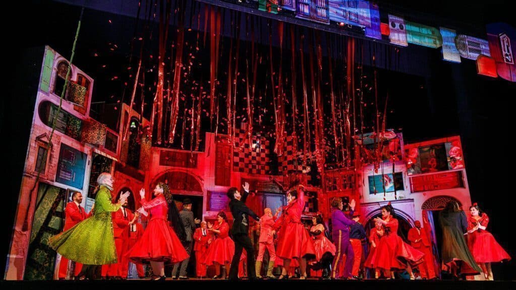 A vibrant stage performance with actors in red costumes, confetti falling from above, and colorful buildings as the backdrop.
