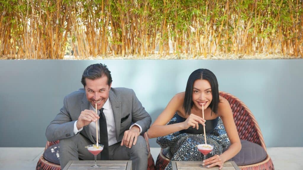 Two people sit on wicker chairs, leaning over small tables and sipping pink drinks through straws, enjoying the finer side of life. A green hedge creates a serene backdrop, making this Anaheim oasis feel like a true retreat.
