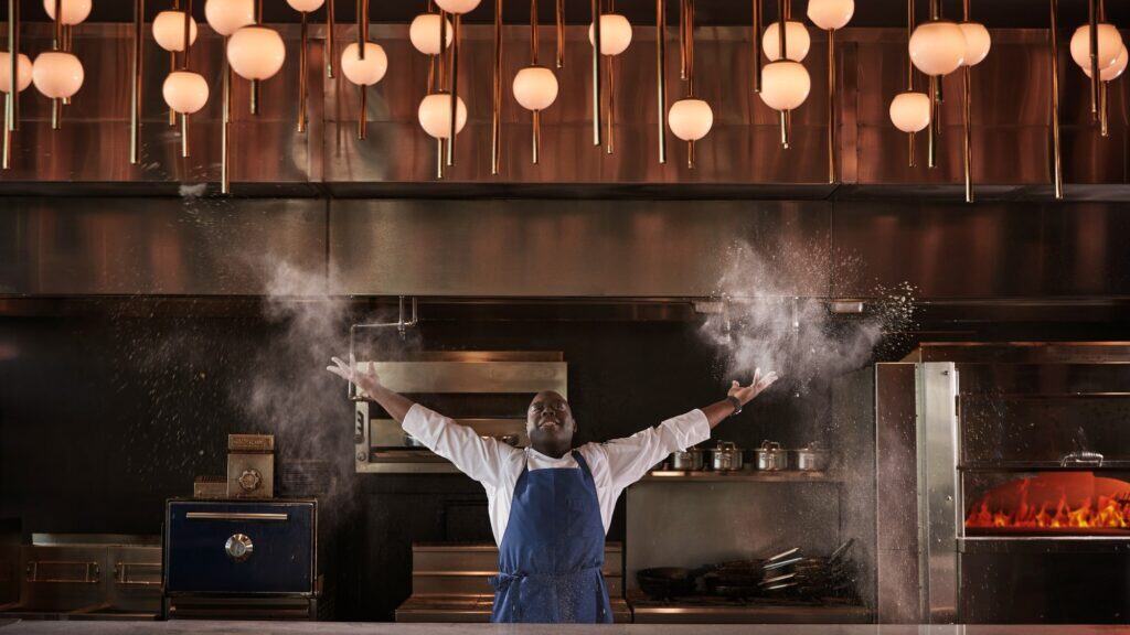 A chef in a blue apron stands in a professional kitchen in Anaheim, throwing flour into the air with both hands under modern round pendant lights, showcasing the finer side of culinary artistry.