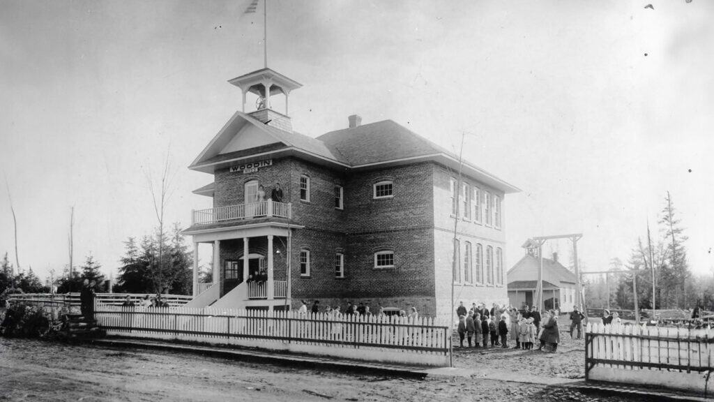 A historical black and white photo showing a group of people gathered in front of the Woodinville Schoolhouse, a two-story brick building with a bell tower, surrounded by a white picket fence.