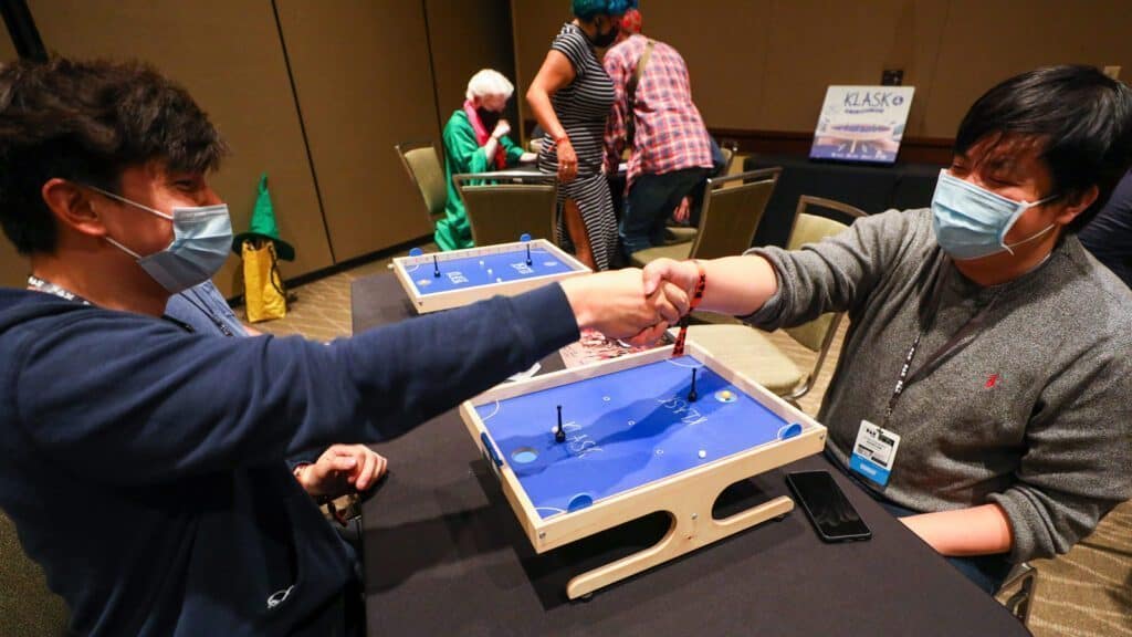 Two people wearing face masks shake hands over a small table game featuring Mario in a room with additional people in the background, also wearing masks and playing games.