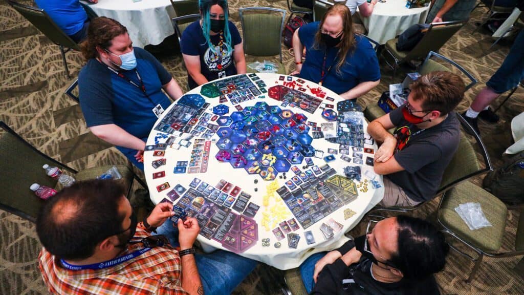 Six people sit around a table in Seattle playing a complex board game with cards, tokens, and tiles spread out. Several players, wearing face masks, excitedly discuss their strategies while a Pikachu plush peeks out from one corner of the room.