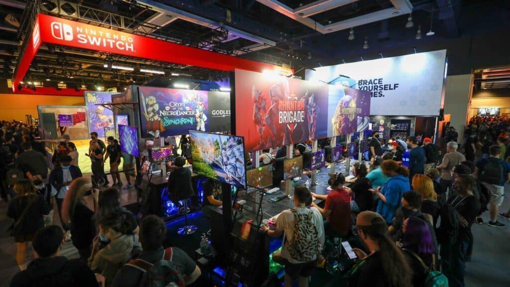 A crowded gaming convention in Seattle with people exploring various gaming booths, including a prominently featured Nintendo Switch area. Multiple screens display different video games, and you can't miss the giant Pikachu and Mario cutouts attracting eager fans.