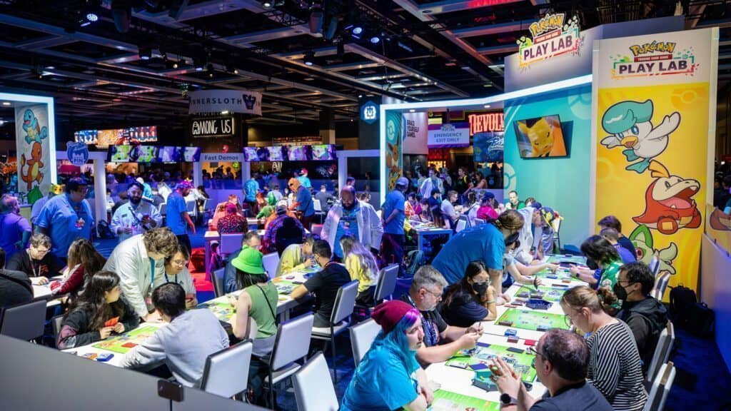 People are gathered at various tables in a convention hall in Seattle, participating in activities related to Pokémon. The setting is vibrant, with colorful posters featuring Pikachu and signs displaying the Pokémon theme.