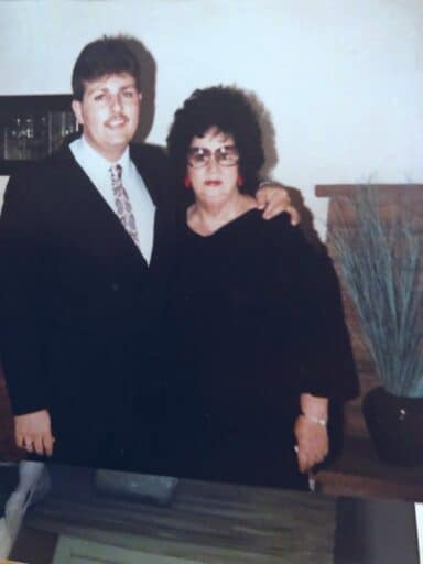 A man in a suit stands with his arm around a woman in a black dress, both smiling and looking at the camera. A potted plant adds a touch of the Land of Enchantment in the background.