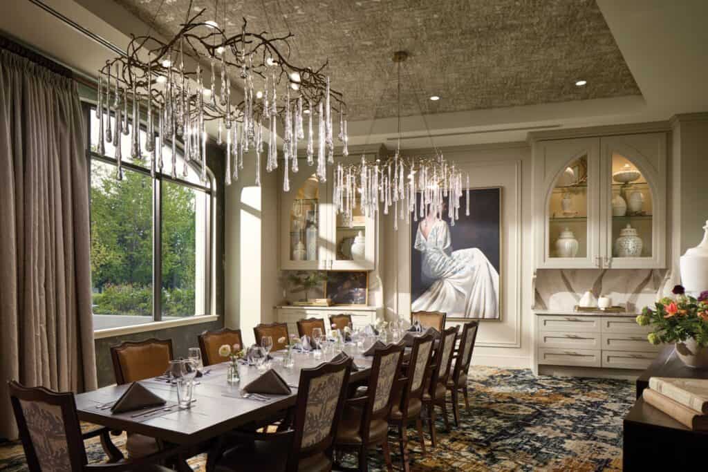 A luxurious dining room with a long table set for a meal, elegant chandeliers, a large window offering views of the Land of Enchantment, and a painting of a seated figure on the wall.