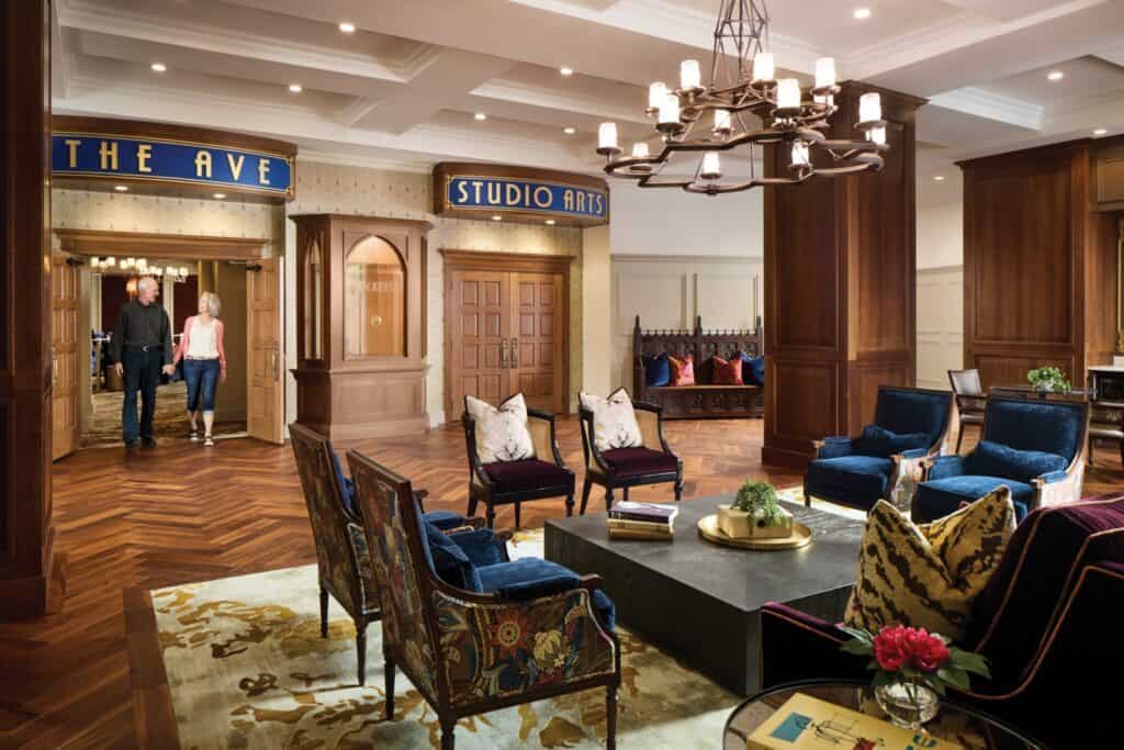 A hotel lobby with wooden flooring, blue upholstered chairs, a chandelier, and decorative cushions evokes the Land Of Enchantment. Two people are entering through double doors labeled "The Ave Studio Arts.