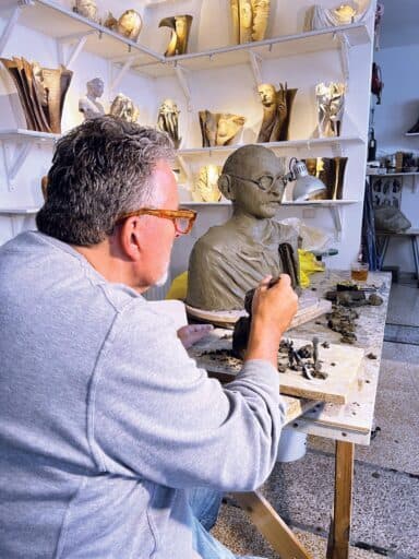 A man with gray hair and glasses sculpts a clay bust in a studio filled with various sculptures and busts on shelves and tables, capturing the magic of the Land of Enchantment in each piece he creates.