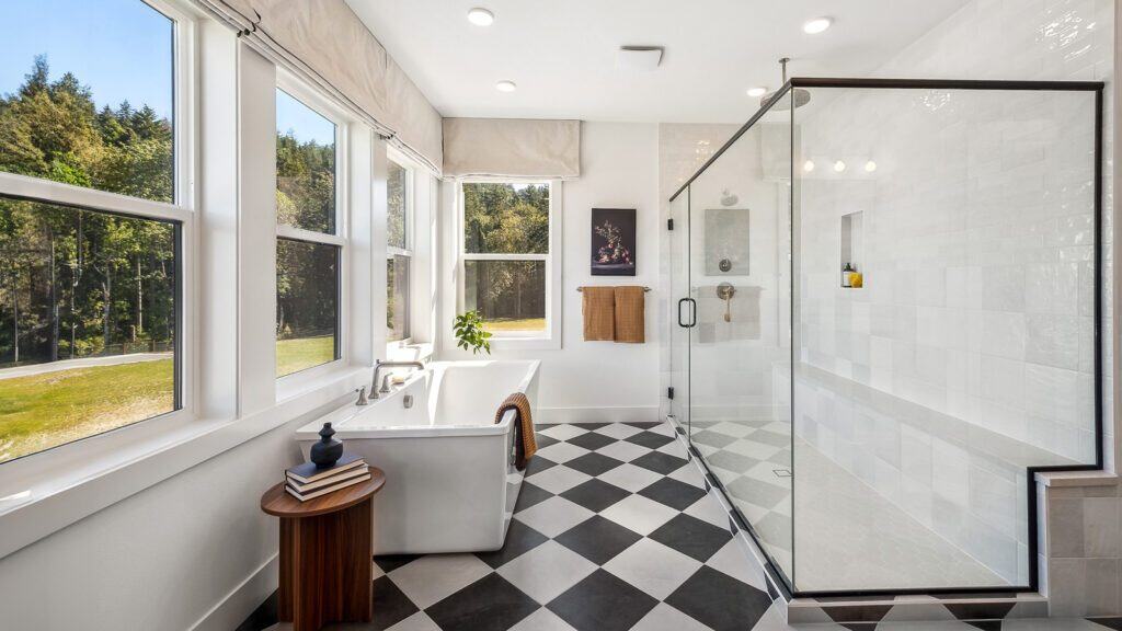 A modern bathroom inspired by Bobby Berk of Queer Eye features a freestanding bathtub, large windows, black and white checkered flooring, a glass-enclosed shower, and a small wooden side table. The space is well-lit with natural light.