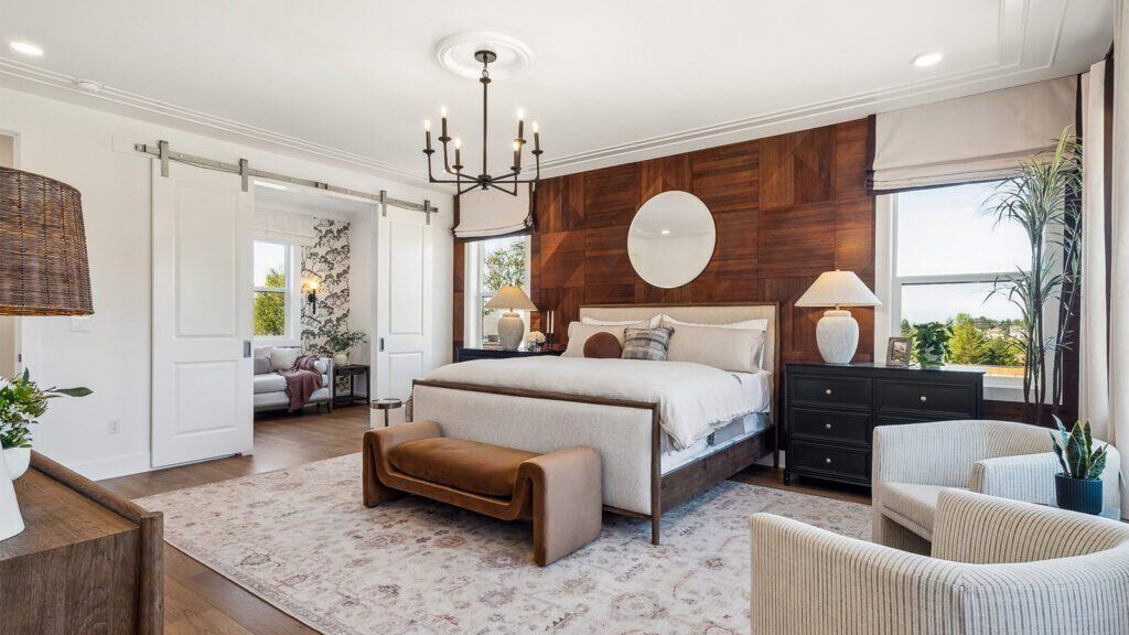 A modern bedroom with a large bed, a wooden accent wall, and a round mirror. Styled by Bobby Berk from Queer Eye, it features a chandelier, carpet, bench, drawers, plants, and a sliding door leading to an adjacent room. Perfect for creating your own Newcastle-inspired sanctuary.
