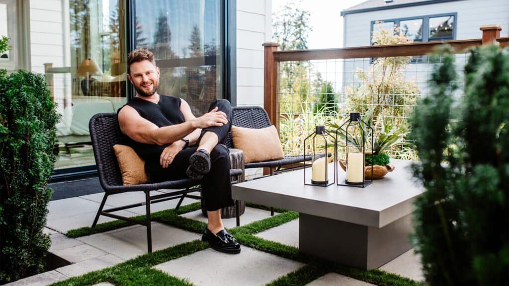 A man dressed in black sits casually on an outdoor patio chair, smiling. The patio, reminiscent of a Queer Eye makeover by Bobby Berk, features a coffee table with decorative lanterns and lush greenery surrounding the seating area.