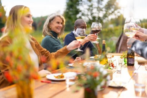 A cozy group of people sits at an outdoor wooden table in Tualatin Valley, Oregon, raising wine glasses in a toast. Various dishes and flowers adorn the table, while the background features trees and greenery.
