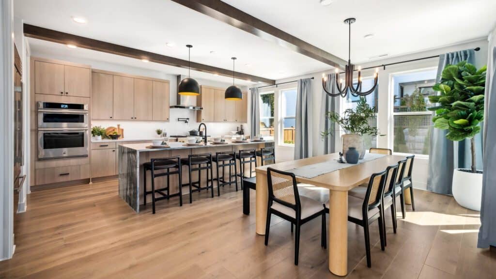 A modern kitchen and dining area, reminiscent of a Bobby Berk design from Queer Eye, features light wood cabinets, a large island with seating, stainless steel appliances, and a chandelier over a wooden table with six chairs.