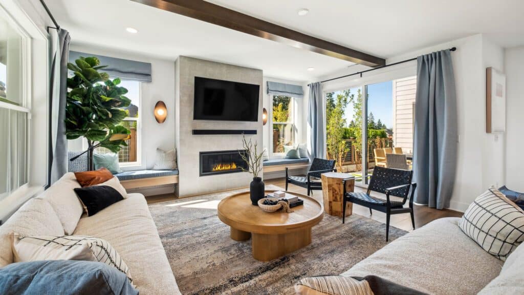 A modern, sunlit living room designed by Bobby Berk features two sofas facing a round wooden coffee table, a large TV above a fireplace, and a sliding door leading to an outdoor deck. This Newcastle-inspired space brings the charm of "Queer Eye" sophistication to your home.