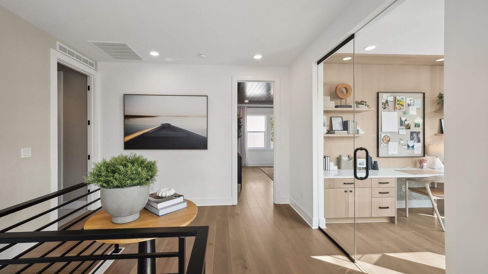 Modern hallway with a small round table holding a potted plant and books on the left, and a glass-fronted office space designed by Bobby Berk of Queer Eye on the right. Doorway leads to another room at the back.