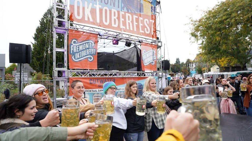 A crowd holds large beer mugs in front of a stage with banners reading "Fremont Oktoberfest" during an outdoor Seattle Oktoberfest celebration on a cloudy day.