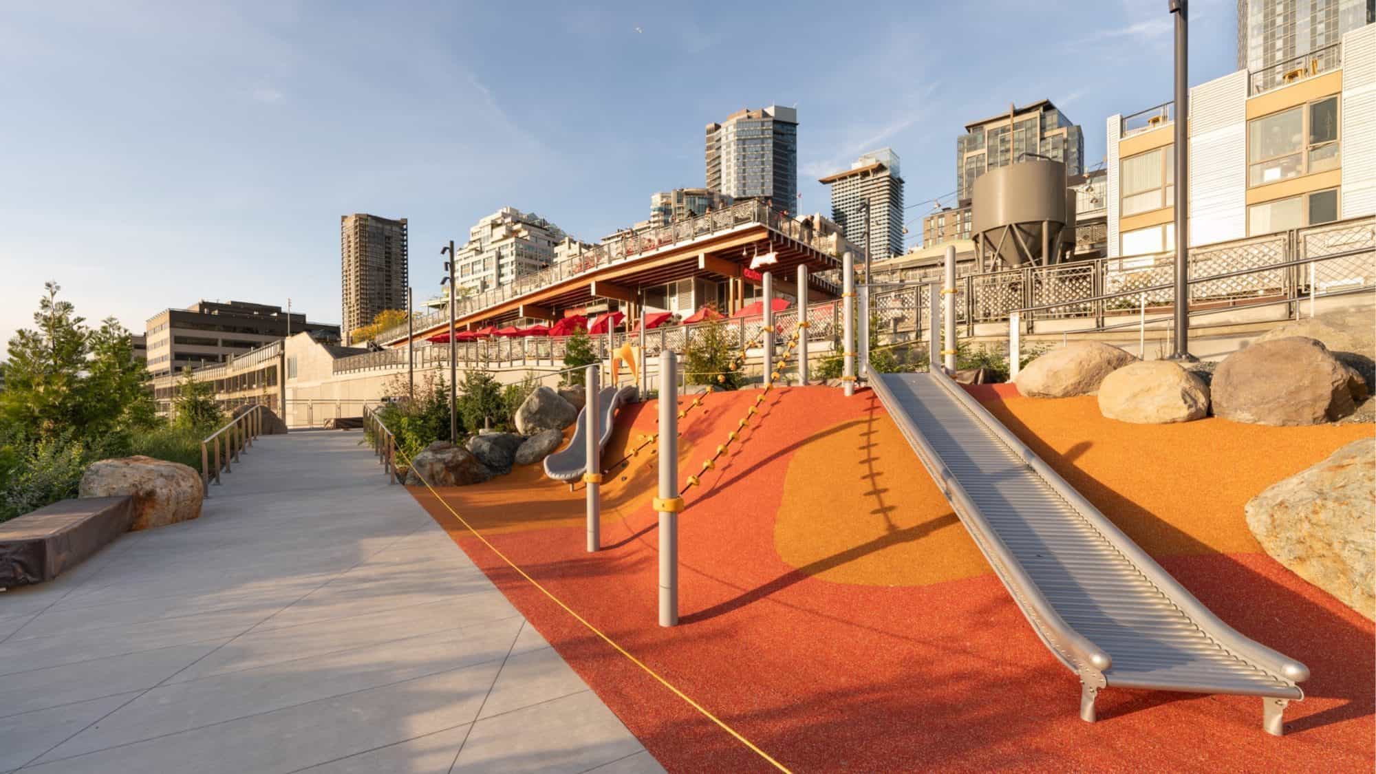 Bluff_Playground_Credit_CityofSeattle-16-9-2000x1125 A transformative playground features slides and climbing structures on vibrant rubber flooring, nestled beside Seattle's Waterfront. Adjacent to Overlook Walk, it offers views of urban walkways and high-rise buildings in the background.