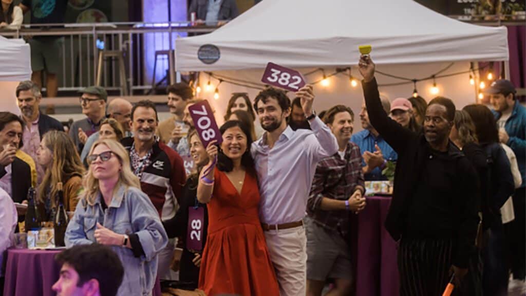 People at an auction event holding numbered paddles, with a crowd in the background under a white tent.