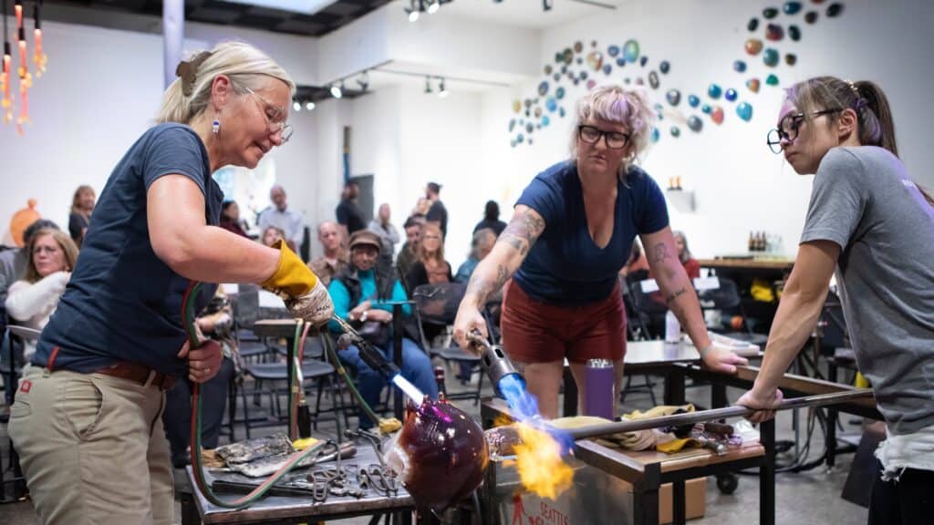 Three people work on a glassblowing project in a workshop, surrounded by onlookers. A woman uses a torch on the glass while others assist.