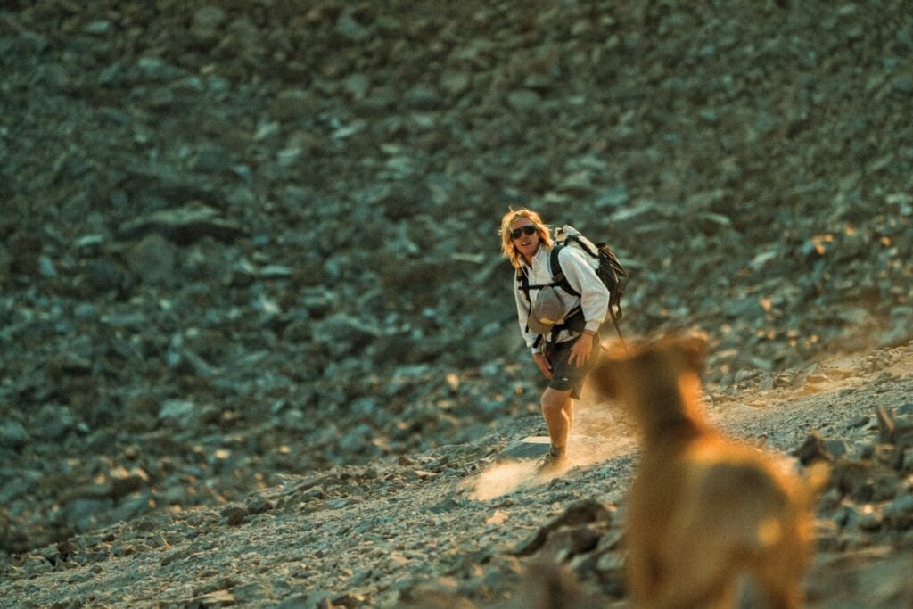 A person wearing a backpack hikes up a rocky slope, quick thinking guiding their path, while a brown dog follows.