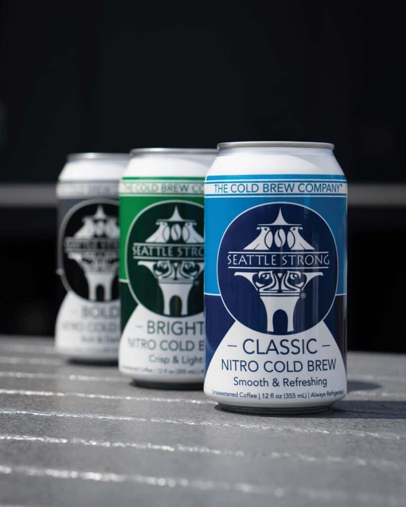 Three cans of Seattle Strong cold brew are lined up in a row on a textured surface: Classic is in the foreground, with Cold Streak and Bold adding depth to the background.