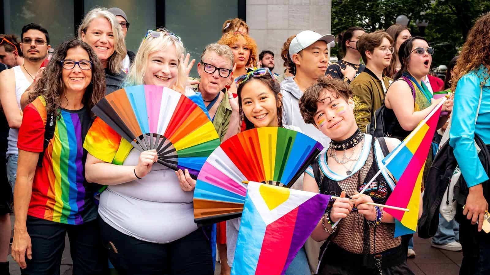 A diverse group of people at Seattle Pride 2025, holding colorful fans and flags, smiling and wearing vibrant clothing, as they help shape a more inclusive future.
