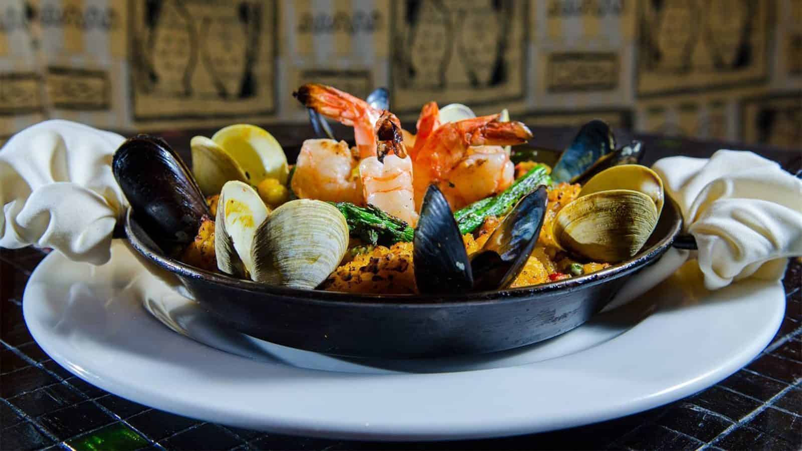 A delightful staycation treat: a seafood dish with shrimp, mussels, and clams on a bed of rice in a black pan, elegantly placed on a white plate with folded cloth napkins beside it.