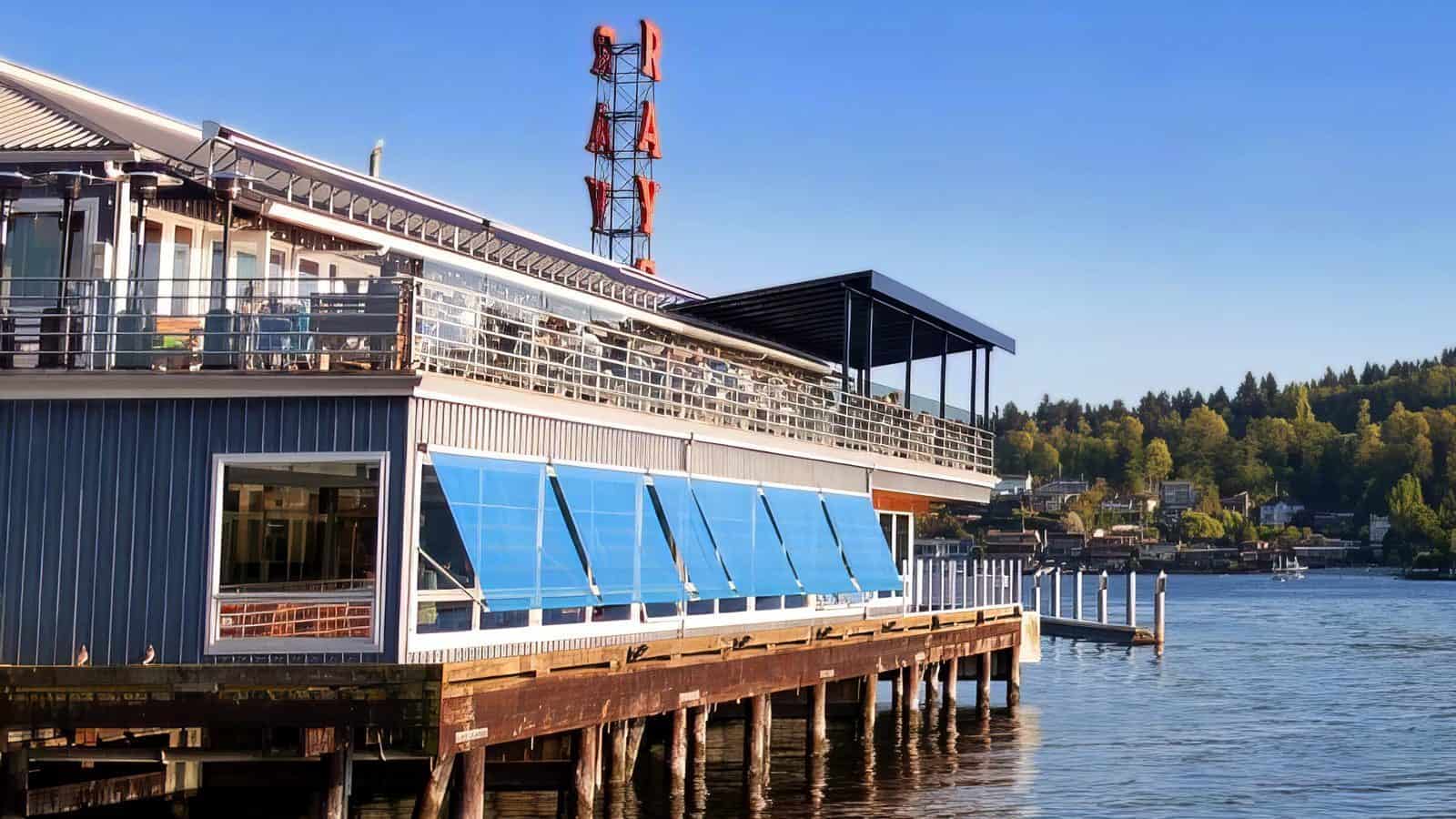 BestOutdoorDining-RaysBoathouse_1600x900 A restaurant with large blue windows on a pier extends over the water, offering outdoor dining with Olympic Mountains views. A neon sign reads "RAY'S." Trees and houses are visible in the background under a clear blue sky.