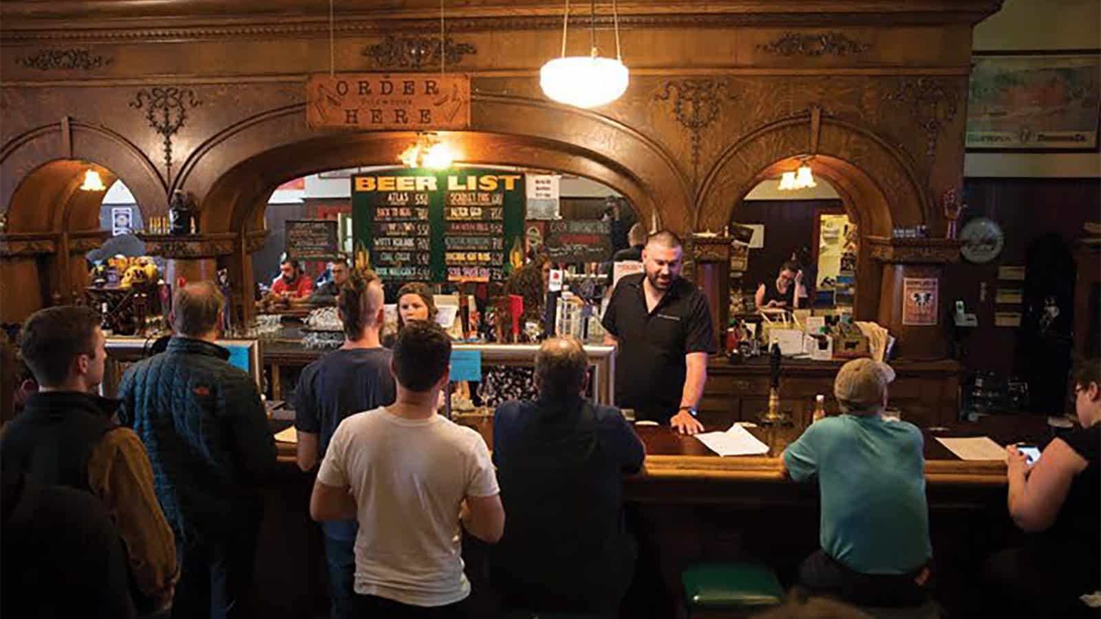 Patrons gather at a historic bar in the U District, where energy hums through the detailed wooden interior. A bartender stands behind the counter, and a large beer list is displayed in the background.