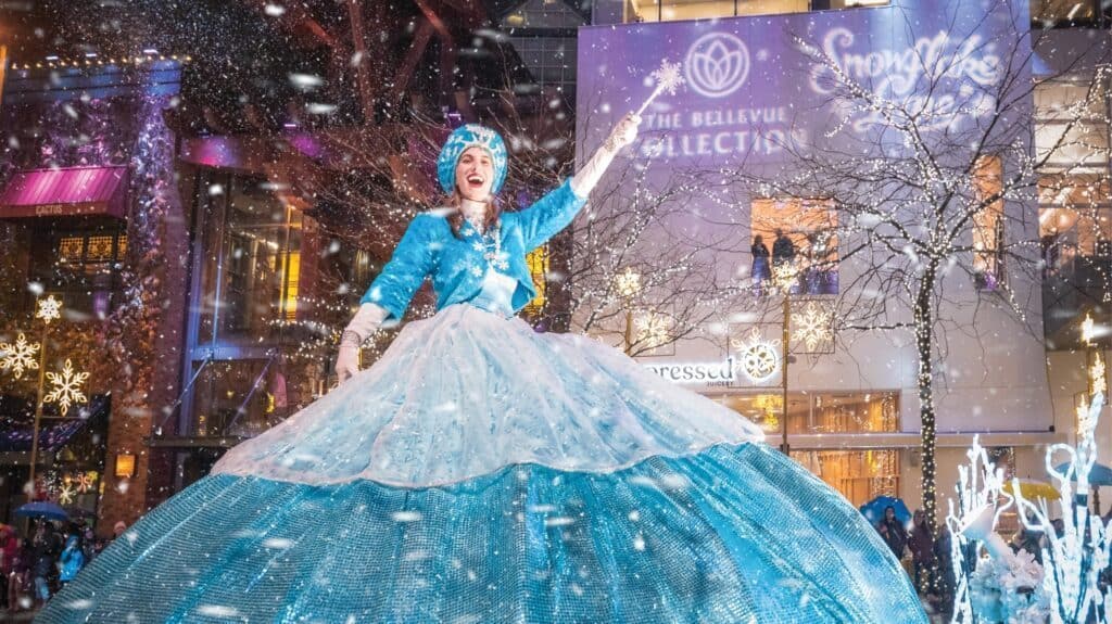 A performer in a blue dress dances outdoors in a snowy setting adorned with festive decorations and a sign reading "Snowflake Lane," showcasing what to do in Seattle during the holidays.