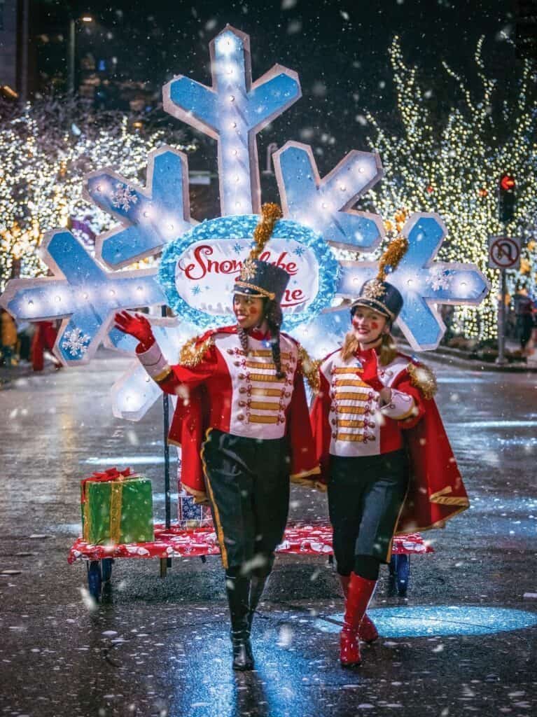Two performers in festive uniforms walk in a holiday parade, smiling under the snowfall. Behind them is a "Snowflake Lane" decoration. For those wondering what to do in Seattle during the holidays, visiting Snowflake Lane and nearby holiday markets and festivals is a must.