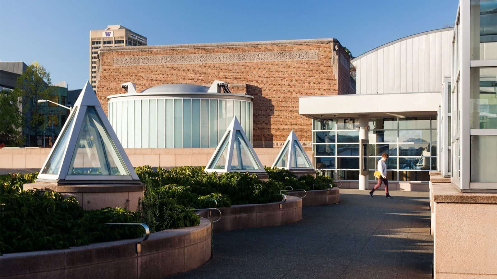 A person strolls through a rooftop garden humming softly, where triangular glass structures glisten, and the energy of the U District buzzes against a brick building in the background.