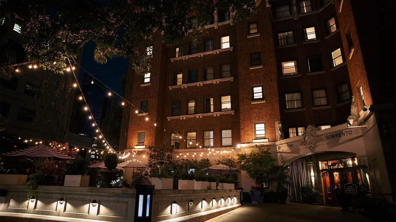 A brick building at night, adorned with string lights and a cozy courtyard with seating, invites you to unwind on your ultimate staycation. The welcoming sign reads "Hotel Sorrento.