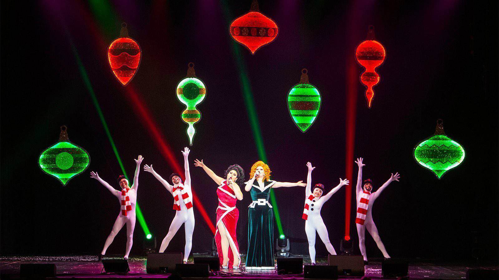 Performers in festive costumes sing and dance on stage, capturing the merry and seasonal spirit while large Christmas ornament decorations hang above.