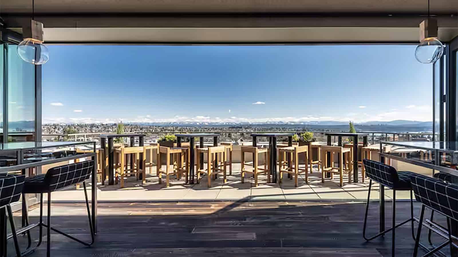 Rooftop bar with wooden tables and chairs overlooking a cityscape, perfect for enjoying under a clear blue sky during your staycation.