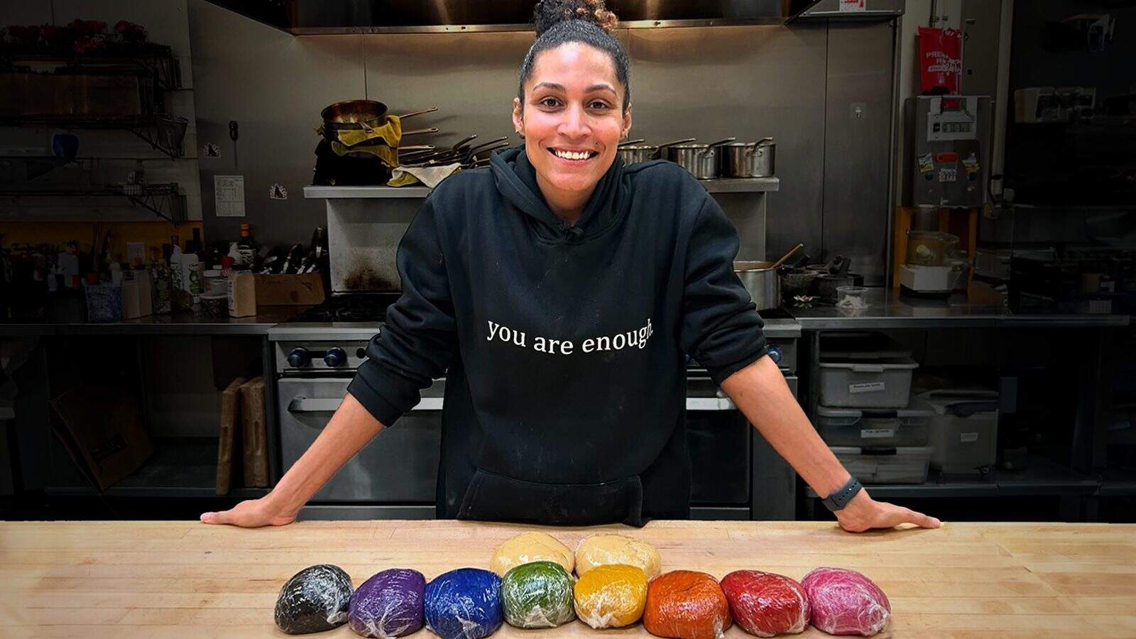 A smiling chef stands in a kitchen behind a wooden table, where eight colorful dough balls are wrapped in plastic wrap, showcasing his creative roots.