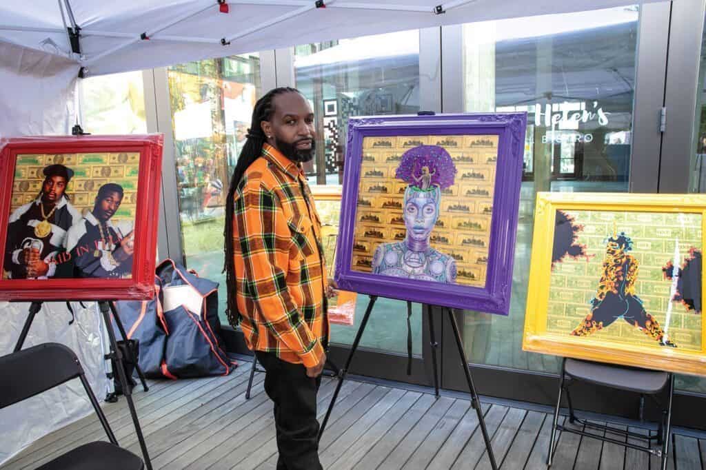 An artist stands beside three colorful framed artworks displayed on easels under a tent, capturing the vibrant spirit often felt in Seattle's hot attractions.
