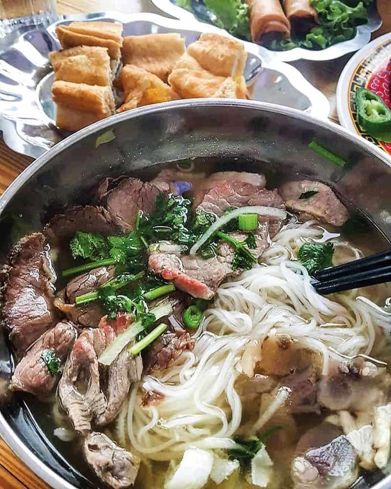 A steaming bowl of beef pho with noodles, herbs, and broth is accompanied by a plate of spring rolls and pastries in the background. This delicious spread is perfect after a day exploring Seattle's hot attraction: the unique and exciting hot tub boat experience.