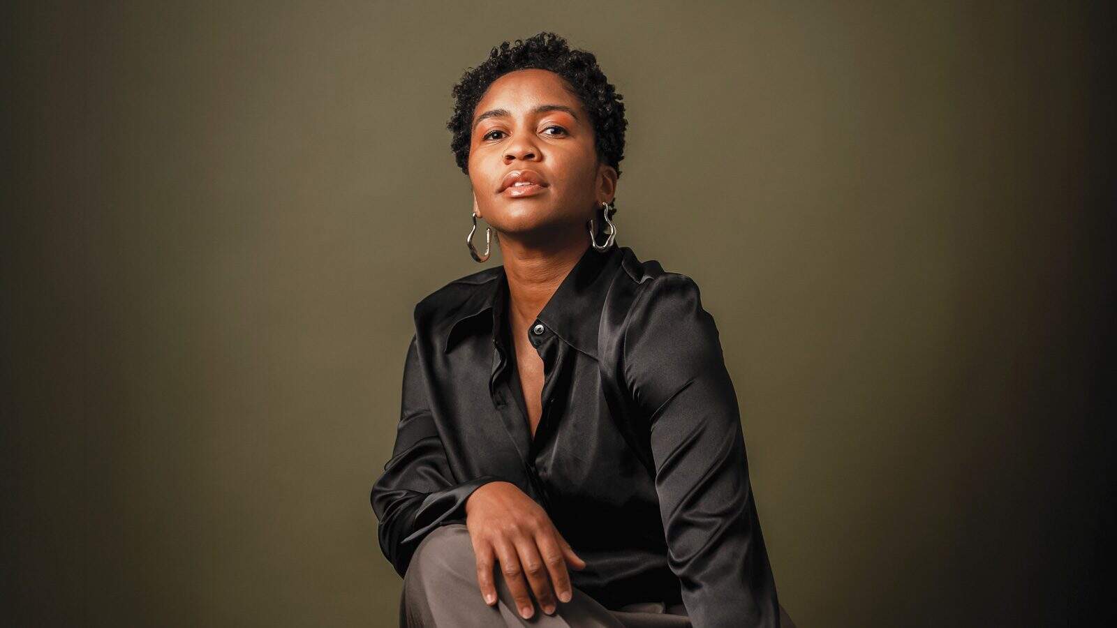 NiaAminaMinor_1600x900 Person in a black shirt, much like dancer and choreographer Nia-Amina Minor, sits with a thoughtful expression against a plain background.