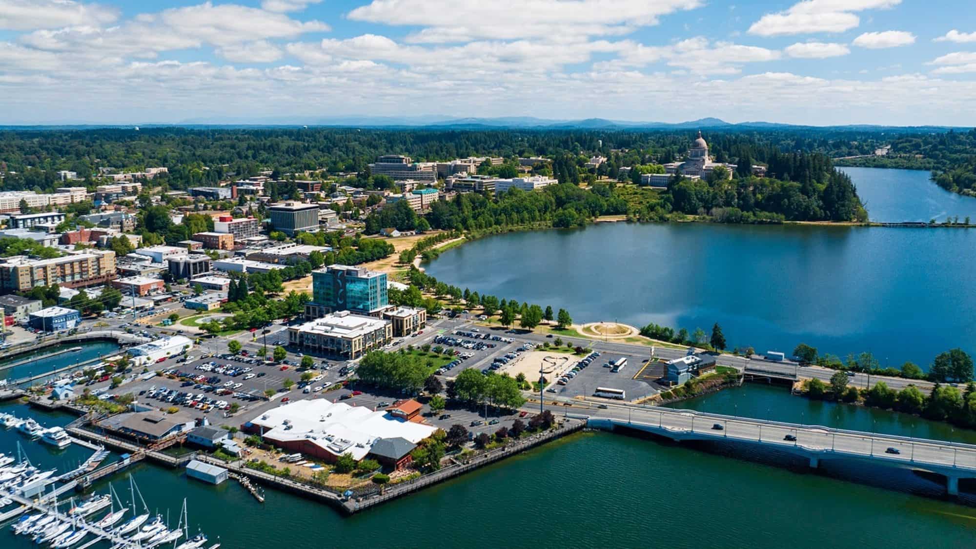 324426216_1979997382344363_7461358672017655969_n-16-9-2000x1125 Aerial view of Olympia showcasing a lake, marina, and bridge surrounded by lush greenery under a partly cloudy sky. The city harmoniously blends nature with innovation from its thriving tech sector.