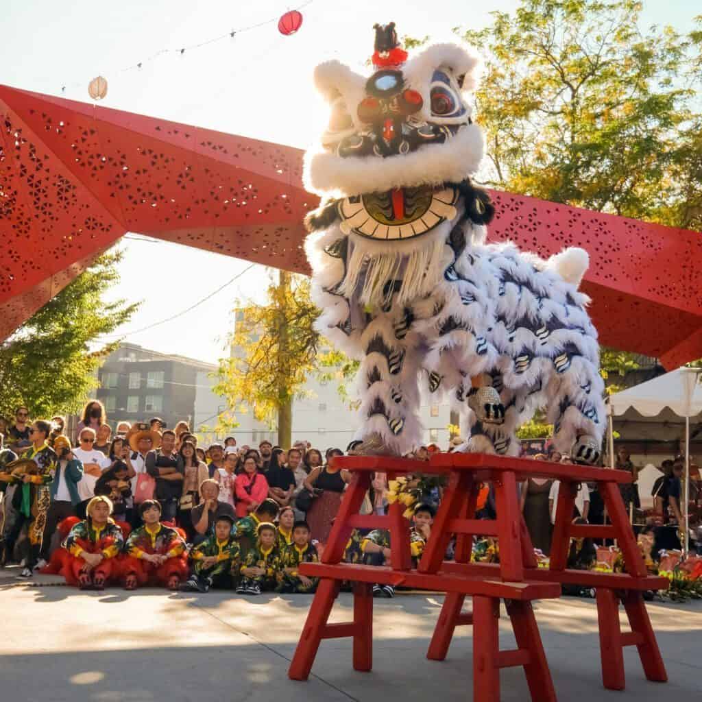 A traditional lion dance unfolds under the light of red lanterns, with a performer on a red bench in a vibrant costume. The gathered crowd watches intently, as occasional raindrops add a touch of drama to this lively outdoor celebration.