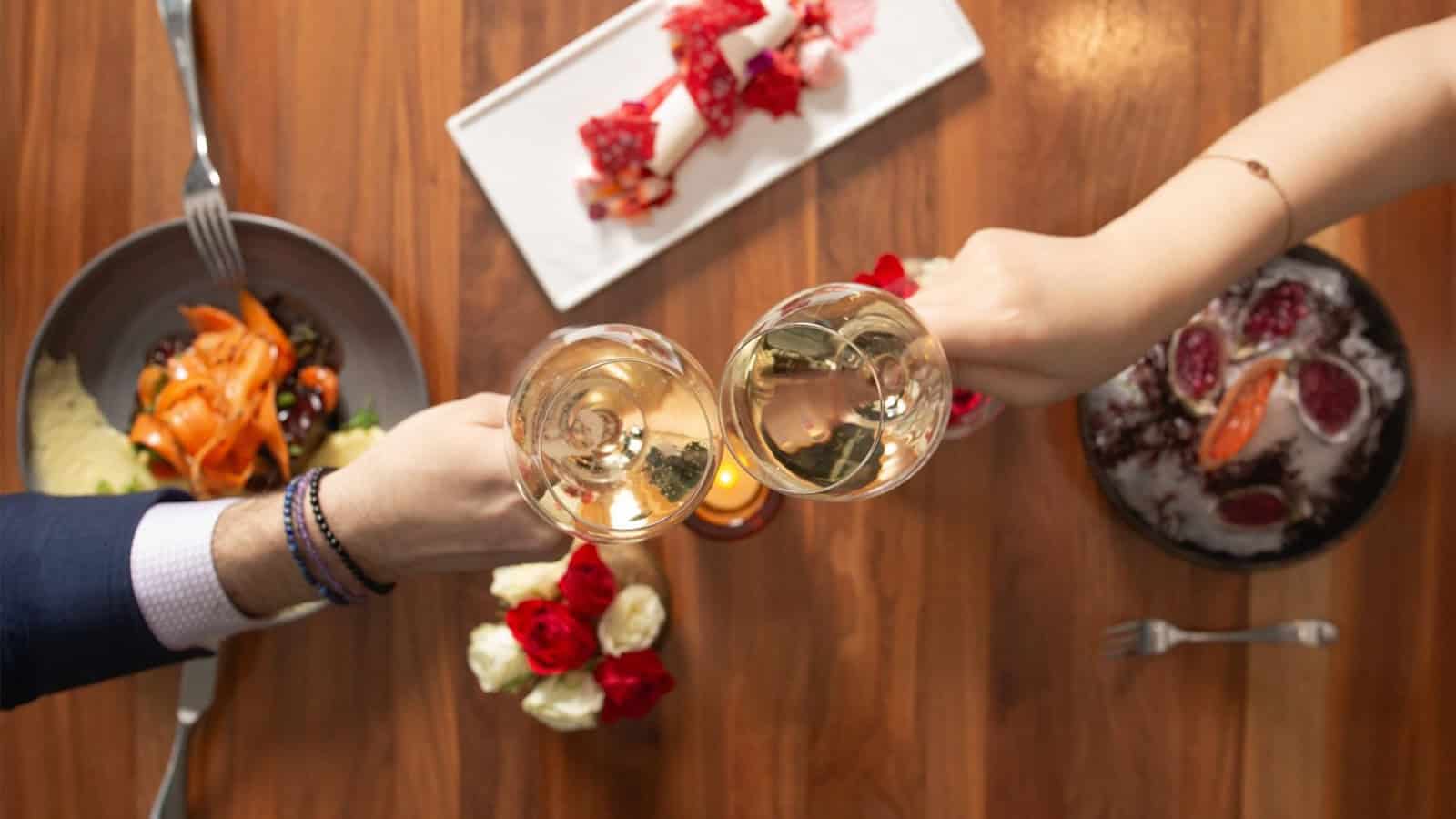 Two people attract champagne glasses on a wooden table, decorated with plates with food and a bouquet of red roses, putting the perfect scene for Valentine's Day to remember.