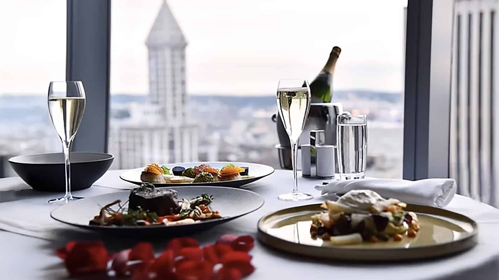 Table to the window in the apartment shows two plates with gourmet food, two glasses of champagne, a bottle of champagne and a set of salt and black pepper. In the background is a city landscape with a pointed tower, perfect for a romantic Valentine's Day.