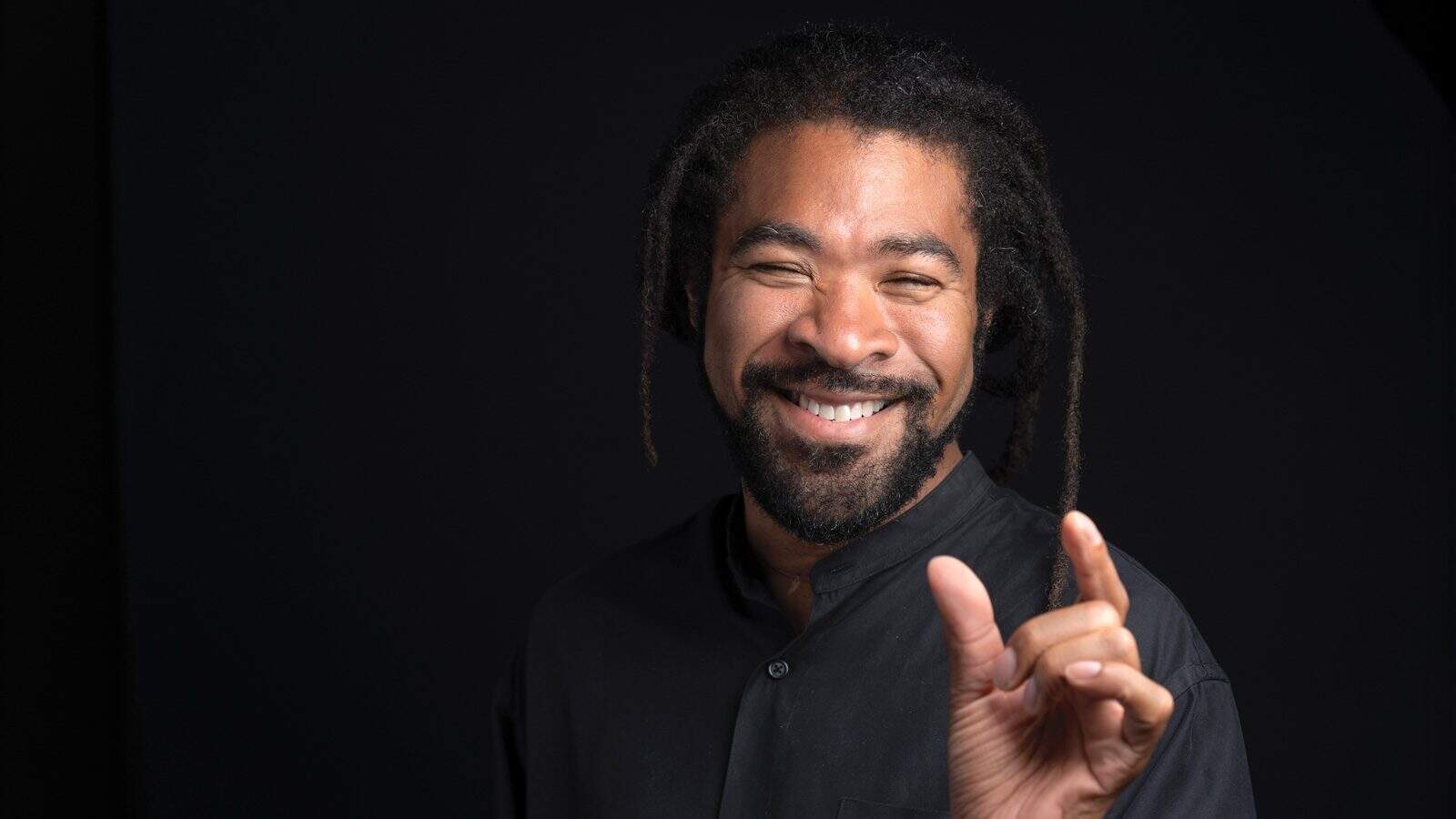 Rico Quirindongo, a smiling man with dreadlocks, holds his fingers apart in a black shirt against a dark background.