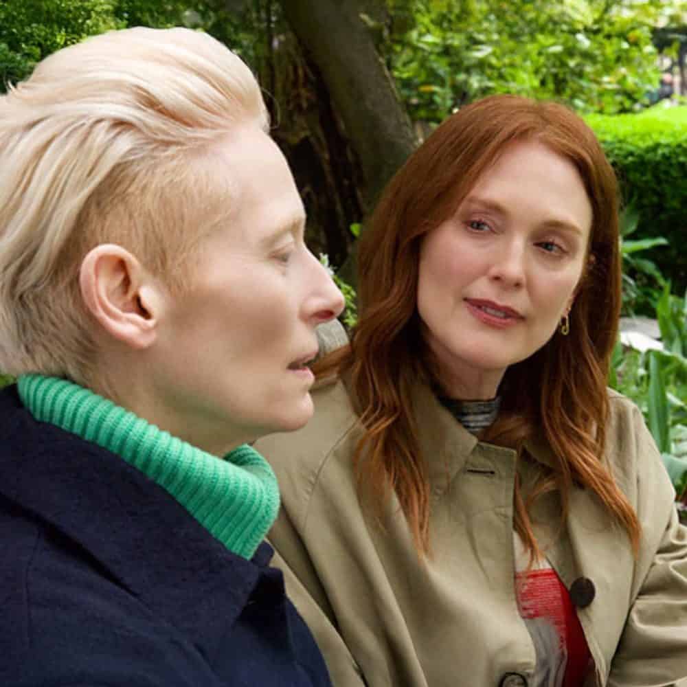 Two women sit outdoors, deeply engaged in conversation. One sports a green turtleneck and short blonde hair, while the other wears a beige coat over her long red hair. Surrounded by lush greenery, this moment captures the essence of a Must List meeting on 01-07-2025.