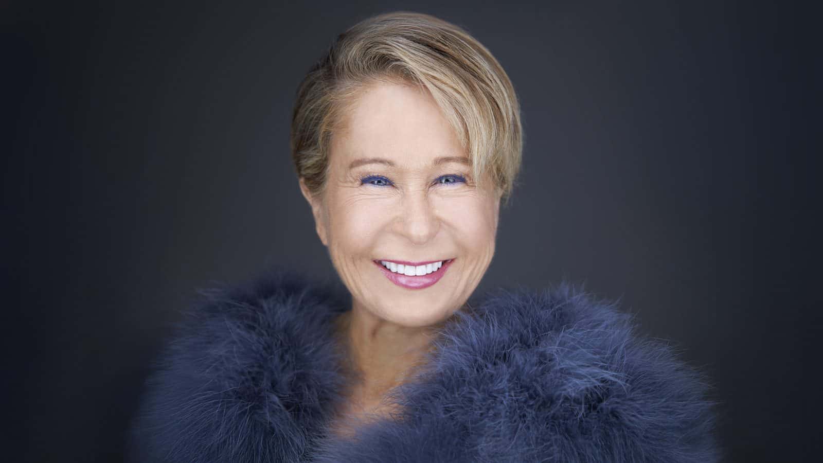 YeardleySmith_1600x900 A person smiling warmly, dressed in a dark fluffy garment, stands against a shadowy backdrop. The scene feels like it could be plucked from the dynamic world of live true crime storytelling, where every detail whispers secrets waiting to be uncovered.