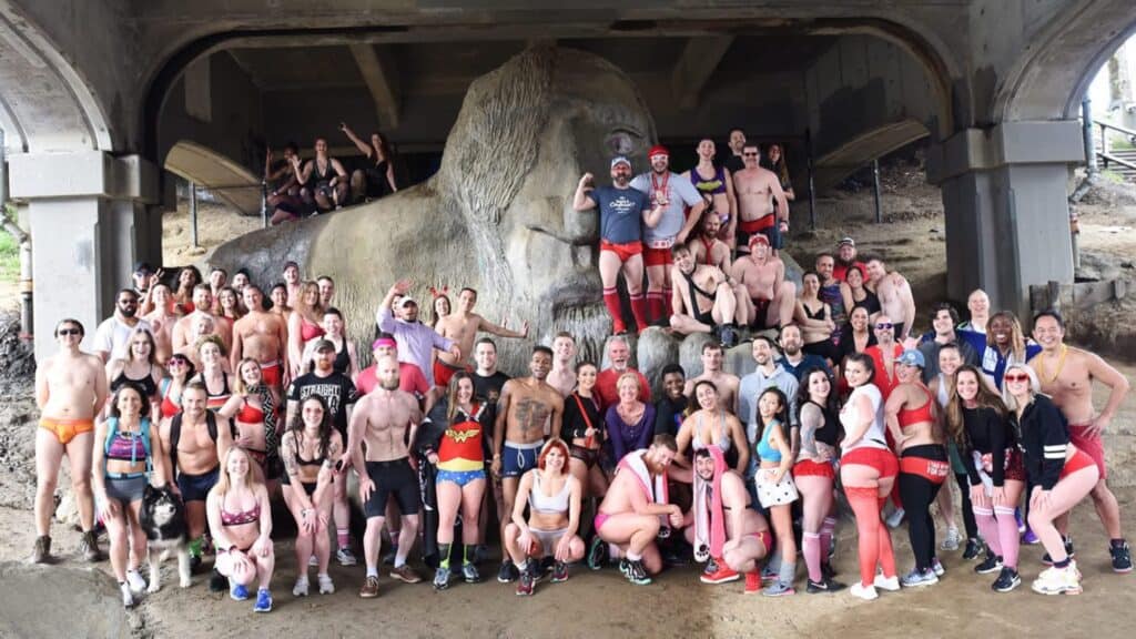 A large group of people involved in "Lost the pants" The cause is posing together under a bridge together, proudly demonstrating their colorful underwear to a large stone sculpture.