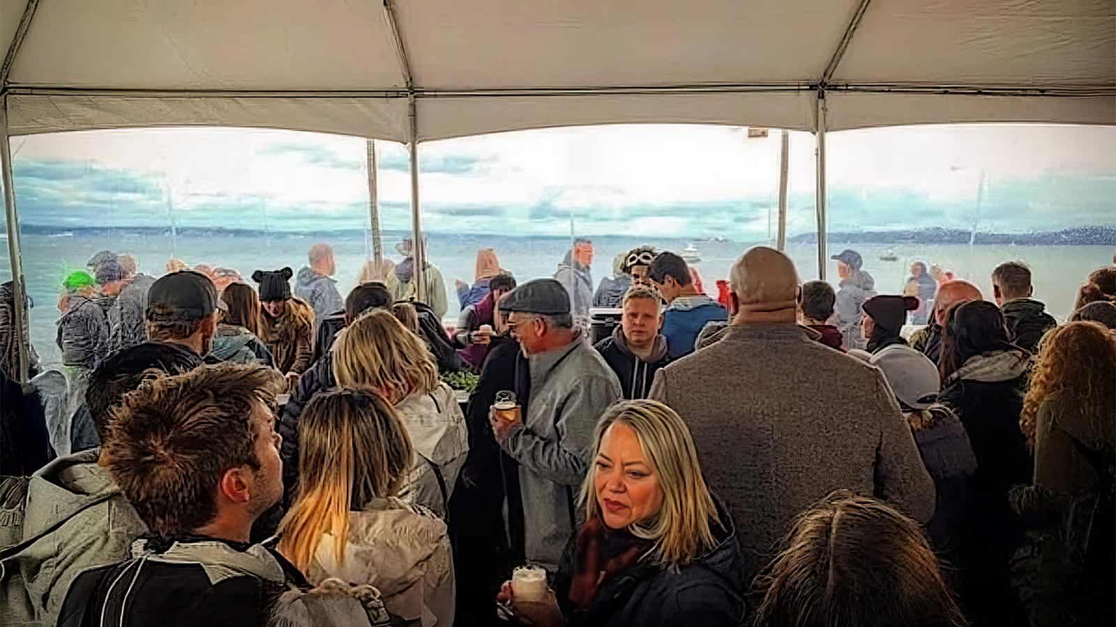 A crowded group of people on a boat deck in Seattle, some holding beverages, enjoys the scenic water view under a cloudy winter sky, all sheltered by a white canopy.