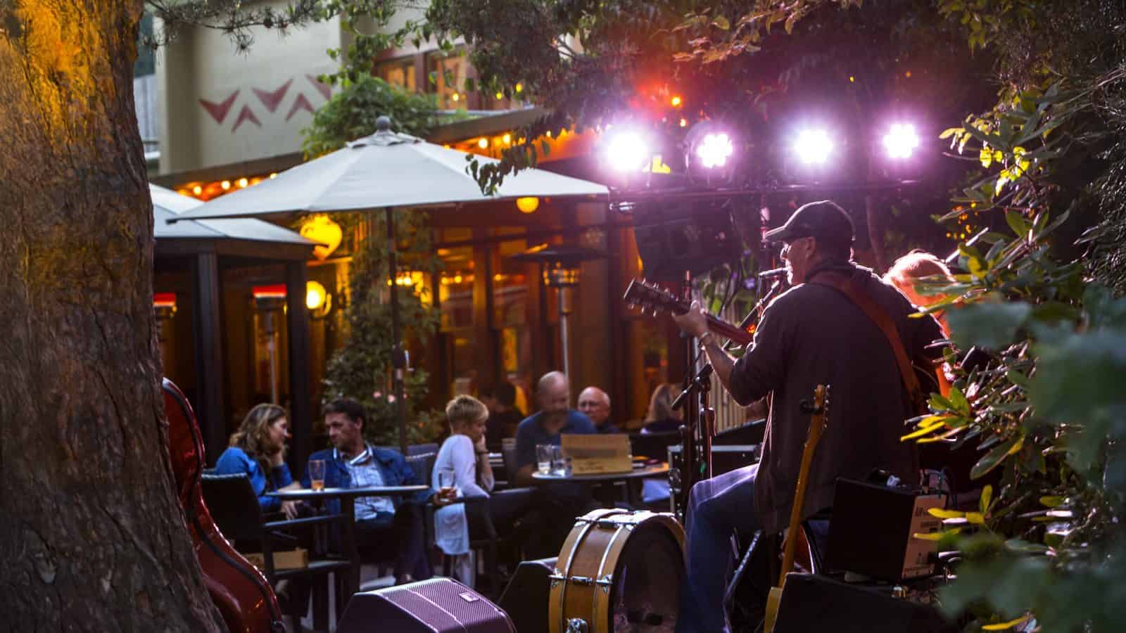 In Seattle’s garden ambiance, musicians perform on an outdoor stage, entertaining an audience seated at tables under patio umbrellas. As part of Seattle's Winter Drinking Plan, guests sip warm beverages, enveloped in a cozy atmosphere perfect for brisk evenings.